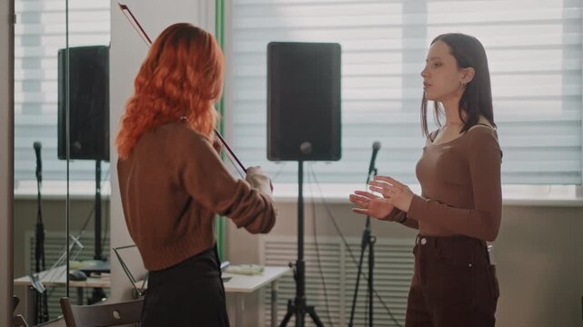 Violin lesson in bright rehearsal room with female coach guiding student through bowing technique, instrument handoff, posture correction, microphone and speaker visible, window light creating warm