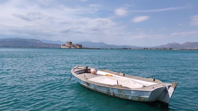 View of a charming traditional Greek fishing boat resting in the turquoise waters of Nafplio Bay, framed by the iconic Bourtzi Castle and the scenic coastline of Pelo