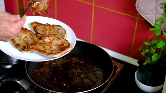 Male hand lifting four cooked pork steaks from frying pan with fork in warm terracotta kitchen