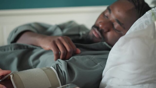 Black patient sleeping in bed at home, closeup of resting face and hand bedside blood pressure cuff and compact monitor on blanket, soft morning light, calm recovery atmosphere, caregiver preparing