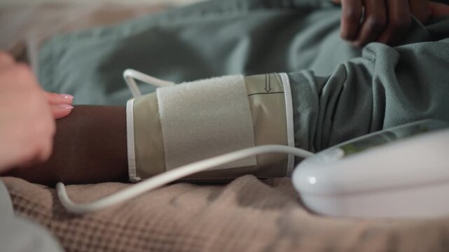 Black man arm with cuff being placed by nurse, closeup of hands securing strap, cable connection to digital monitor, careful touch, home bedding background, clinical yet comforting action