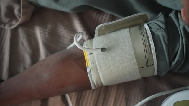 Black man upper arm with blood pressure cuff secured by paramedic, velcro strap and tubing in view, steady positioning for measurement, home bedding, close inspection and calm clinical focus