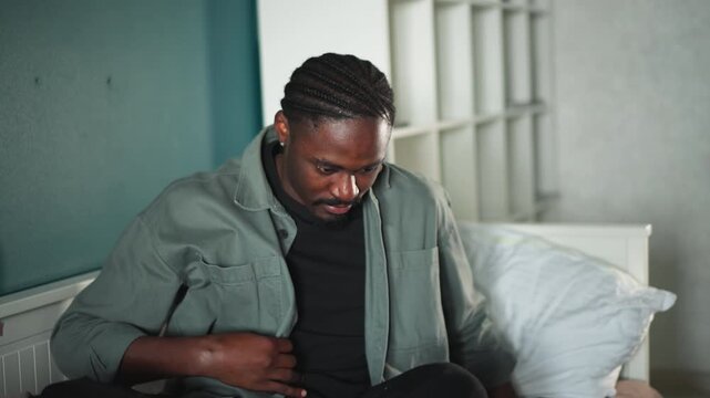 Black man patient lying on bed in dim bedroom, unsteady movements, reaching toward bedside, tired expression, casual shirt, soft pillow, quiet tension suggesting recent collapse or lightheadedness,