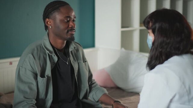 Male patient reveals chest to doctor in home during calm consultation, clinician wearing mask, seated on bed, discussing symptoms, checking vitals and offering reassurance, note taking and gentle