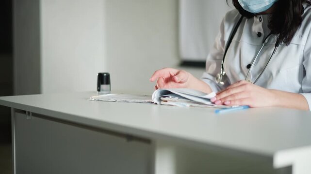 White doctor at desk documenting consultation notes, masked physician reviews chart and writes observations, stethoscope resting on table, clinical office lighting, attentive posture and careful hand
