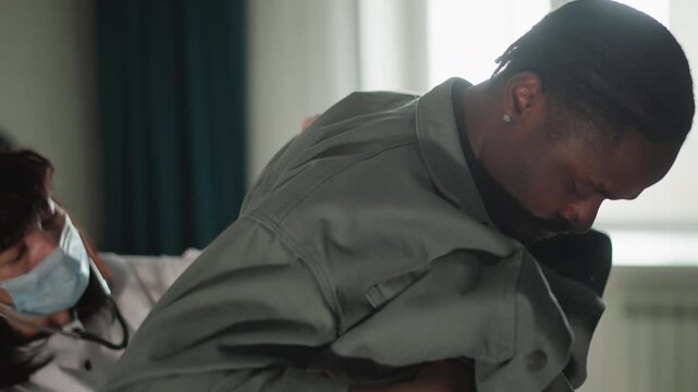 Black man assisted by masked nurse during home check, nurse lifts jacket and assesses shoulder, stethoscope visible, clinical mask and gloves, muted daylight through window, bedside furniture