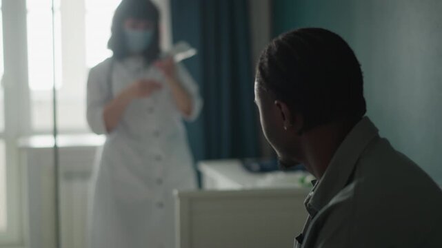 Black patient in clinic with asian nurse preparing medication near window masked nurse measures dose at bedside while seated patient looks on, muted color palette, soft backlight through curtains,