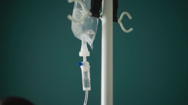 Iv bag hanging against teal wall, slow drip through clear tubing and visible drip chamber on white pole, clinical silence and empty bedside suggest patient offframe, sterile atmosphere ideal