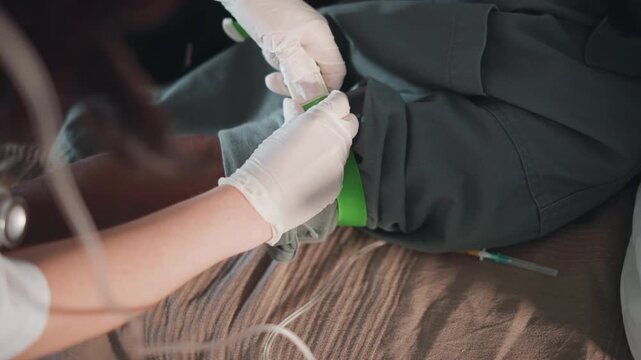 Black patient leg receiving tourniquet during home emergency, paramedic tightening green strap with gloved hands, closeup on wound area and IV tubing, bedside stabilization with calm professional