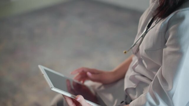 Caucasian doctor thoughtful with tablet in quiet room, clinical contemplation. Stethoscope visible, mask lowered, focused expression while tapping screen and studying records on lap, subdued interior