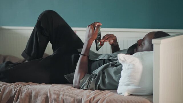 Black man lying on bed scrolling phone for telehealth consult, messaging nurse, awaiting paramedic or ambulance, monitoring symptoms while resting on pillow and blanket in cozy bedroom, calm