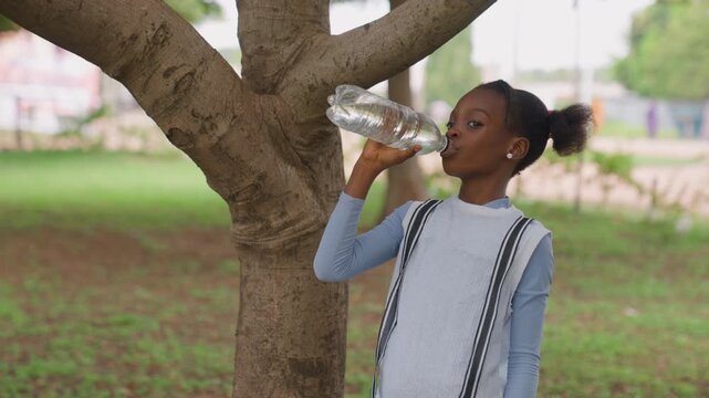 Girl refreshes outdoors. Child enjoys hydration beneath leafy canopy. Serene young girl pauses for water in lush park shade. Peaceful moment as young girl hydrates under tree in quiet park