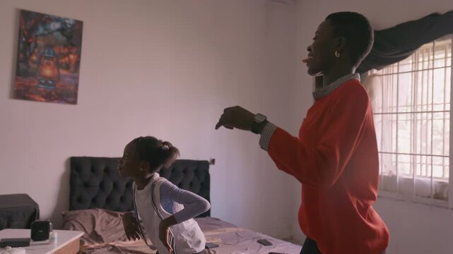 Black mother and daughter dancing in bedroom, playful teaching moment with mother as dance coach and daughter copying steps, sunlight through window, bed and poster in background, casual red top