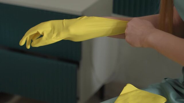 White woman putting on yellow gloves, sitting in living room, sliding rubber gloves over fingers, textured grip visible, calm methodical movements, soft natural light, vintage dress, closeup household