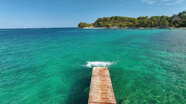 Aerial top view of a young couple jumping from a wooden pier into the clear turquoise sea water. Scenic drone video of people enjoying summer vacation travel.