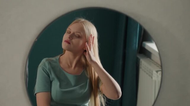 White girl house mirror evening prep glow fixing hair smoothing shirt patting face adjusting necklace poised confident expression soft interior lighting bedroom radiator teal curtain vlogger fashion