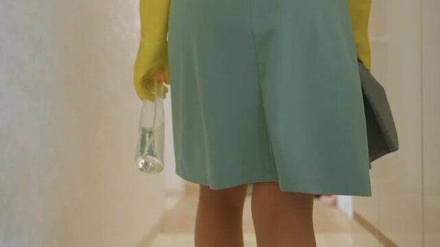 Empty hallway after cleaning routine with closed door and coat hooks, soft light on laminate floor, mop and bucket near threshold, woman walking away with cloth and spray, calm finished task mood,