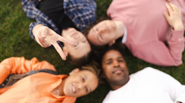 Four people lying on grass, one making a peace sign, blurred focus.