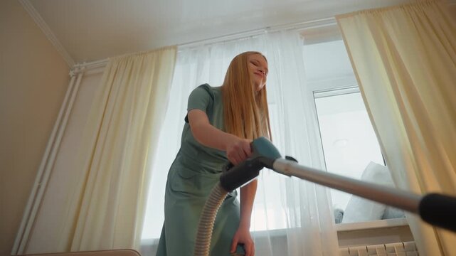 Young white woman vacuuming by window in bright living room. Bright curtains and mint dress frame energetic motion as cleaner persona tidies carpet near sofa and pillows. Lowangle perspective captures