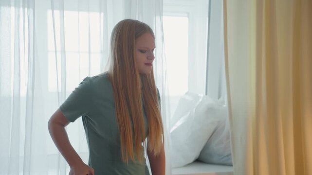Young woman house cleaning by window, long hair and green dress vacuuming near sheer curtain and sofa pillows in soft morning light calm expression, routine domestic chore, parquet floor, cordless