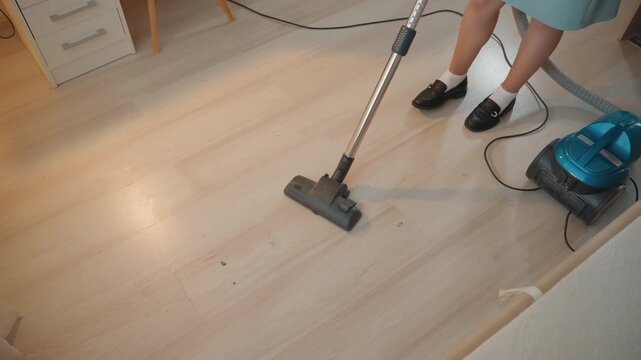 Woman vacuuming light hardwood floor in bright bedroom, turquoise canister vacuum with visible cord pulls along edge while feet in black shoes move across planks closeup of nozzle gliding, dust