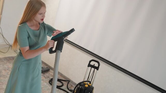 Caucasian woman wrestling carpet vacuum cord. Living room scene with canister machine, hose tangling, determined efforts, frustrated expression, hands gripping handle, pulling and pushing motions,