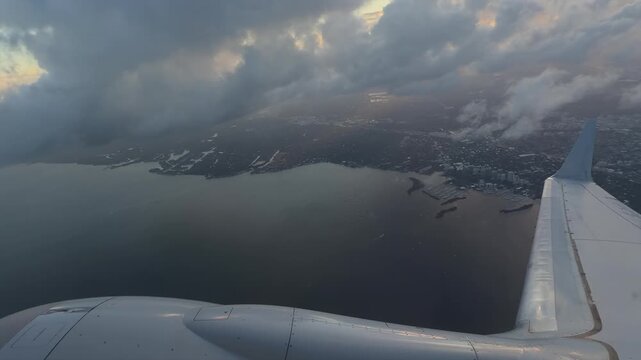 airplane wing and engine flying above city coastline and bay through clouds at sunset