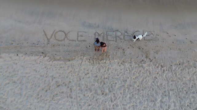 A young couple and a dog on a Brazilian beach with an inscription marked in the sand that says "voce merece" (you deserve) and some beach chairs.