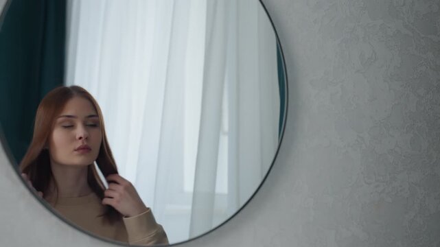 Young woman styling hair in round mirror, steady gaze and practiced movements by sheer curtain, modern bedroom backdrop with soft natural light, decisive preparation and posture suggest readying