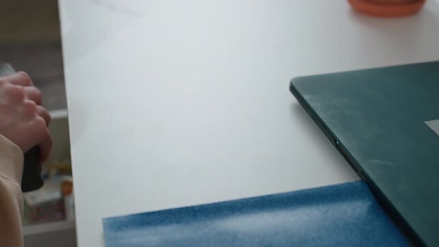 Closeup desk notebook spray bottle hand reaches for eraser pencil green folder on table with blue mat tidy workspace for home tutor preparing grammar exercises soft natural light calm focused
