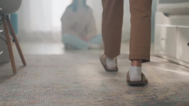 Tutor approaches barefoot and hands grammar book to seated student by window, intimate home coaching scene with carpeted steps, slippers, gentle guidance and supportive mentorship for study