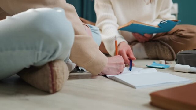 Closeup hands and legs writing caucasian tutor and student seated on floor with notebook and clipboard, pen marks on paper, cozy rug and soft sweaters, warm natural light, quiet grammar coaching