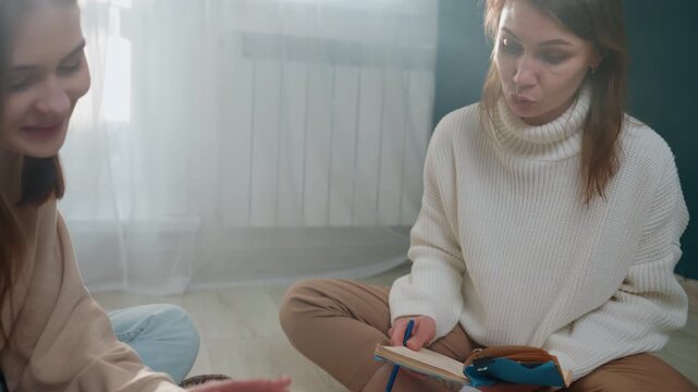 Two women sitting on floor studying together with notebook and pencil, tutor guiding student through notes while tape dispenser and stationery sit nearby soft window light and neutral rug create