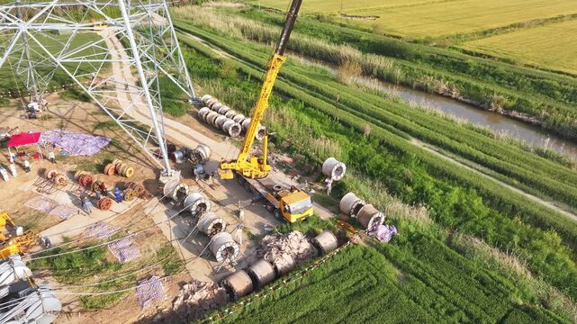 High voltage transmission tower construction site with crane lifting operations in green fields