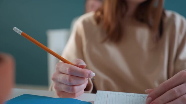 Closeup hands writing math notes on paper grid with pencil and blue folder, beige sweater sleeve visible, methodical strokes and page turning, steady rhythm and concentrated exam preparation