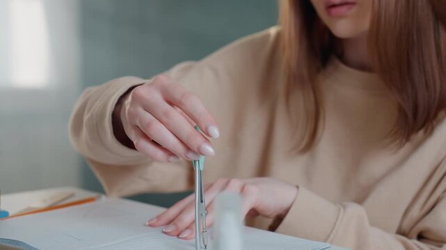 Young white woman using compass on dotted workbook making precise circle measurements during focused home study session wearing beige sweater under soft daylight closeup of hands and compass near pen