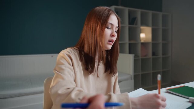 Young woman writing at desk during athome grammar lesson, student fills notebook while tutor gestures with pen, lamplit study room, shelves in background, focused concentration, revision