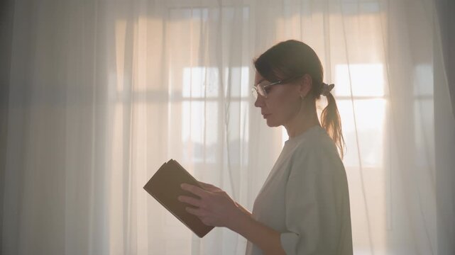 Caucasian woman home reading book, private grammar tutor preparing lesson in backlit room with sheer curtains, soft morning sunlight casting warm glow, reflective profile flipping pages