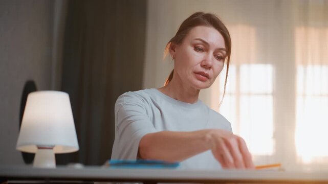 Woman at desk reviewing notes under lamp, flipping book, careful markings, lamplight glow, serious expression, grammar corrections, tutor prepping lesson materials, neat workspace, study mood