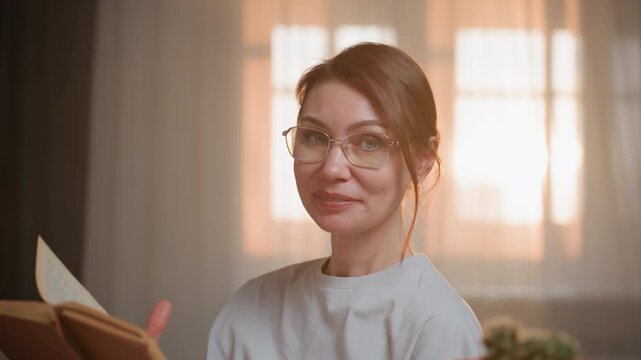 Woman coaching english grammar via video, turning pages with gentle smile, glasses catching warm window light, closeup shot of coach delivering feedback and pronunciation tips, relaxed home lesson