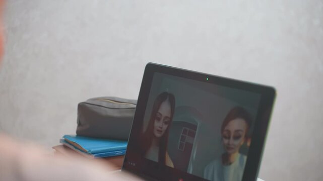 Laptop video call white students on screen viewed from behind, remote grammar session with gestures toward camera, stack of notebooks and phone beside laptop, soft indoor light, attentive faces