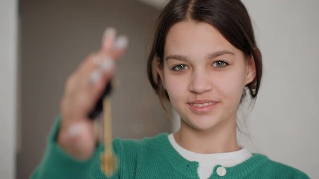 Caucasian teenage woman extending keys towards camera in bright apartment doorway smiling confident green sweater closeup of dangling keys celebrating movein proud new homeowner warm shallow depth