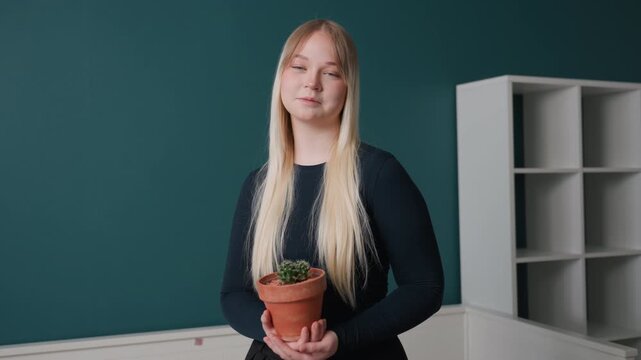 White woman holding plant in apartment hopeful newcomer arranges terracotta pot by empty shelving, teal wall background, long blonde hair, casual black sweater, soft daylight, minimal interior,
