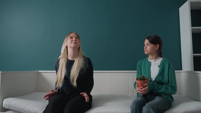 Two women testing daybed comfort in empty apartment while deciding layout, playful gestures, pointing at wall, pot plant between them, unpacked boxes nearby, casual wardrobe, negotiation