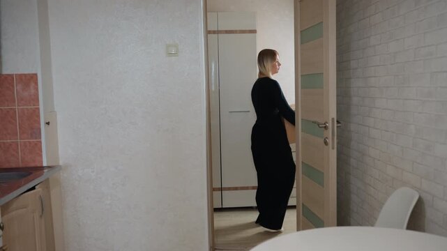 Caucasian woman carrying box into apartment, walking through small kitchen and hallway, opening door, pausing to check layout, placing box near cupboard, portraying renter and unpacker in quiet modern