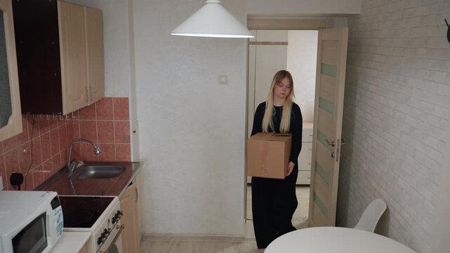 Young caucasian woman carrying moving box into kitchen, entering small apartment through doorway, placing box on round table, pausing to unseal flap and inspect contents, compact cabinets