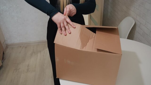 Young caucasian woman opening moving box, carefully unfolding cloth and revealing kitchenware inside cardboard, hands smoothing flaps, closeup of personal items on small apartment table, gentle