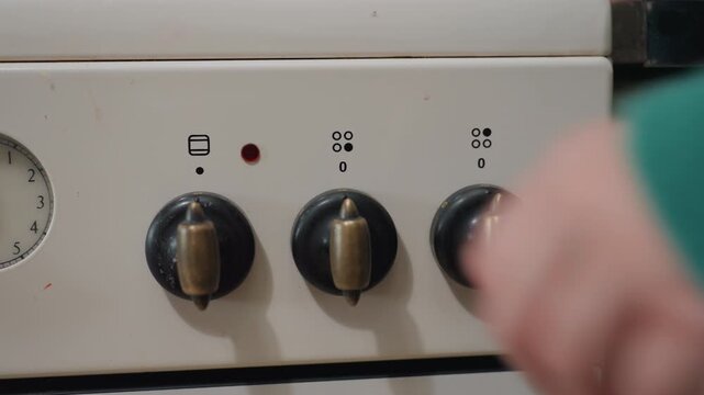 Moving day quick stove safety check, closeup of white range control panel with red power indicator lit, three black brass knobs, pressure gauge visible, green sweater hand turning dial, compact