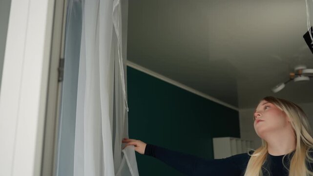 Caucasian woman adjusts curtains new apartment pulls back sheer and drape inspects ceiling fixture and dresser placement empty room with teal accent wall arranges layout tests light and placement