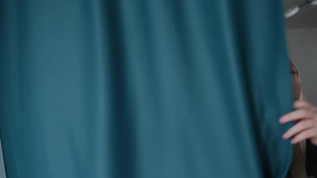 Blonde white woman peeking behind teal curtains exploring empty apartment, assessing layout, imagining furniture placement and natural light, testing fabric texture, thoughtful expression